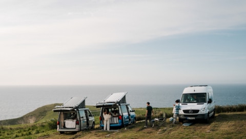 Three campervans parked at the edge of a cliff with people standing around them.