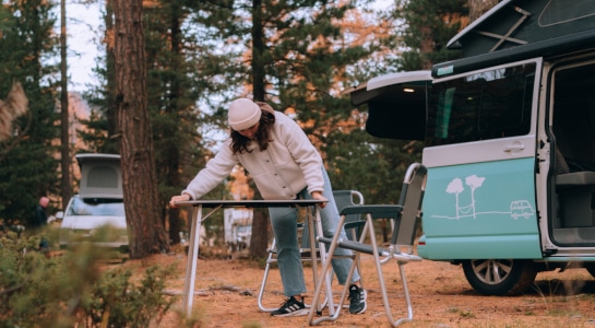 Woman sets up a camping table and chairs at a camping spot where a VW campervan is parked