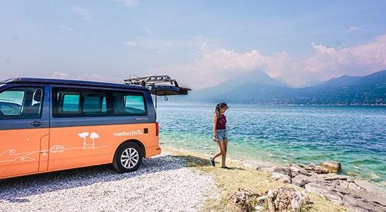 Italy, Lake Garda with a parked roadsurfer camper van, and a girl walking towards the turquoise water of the lake