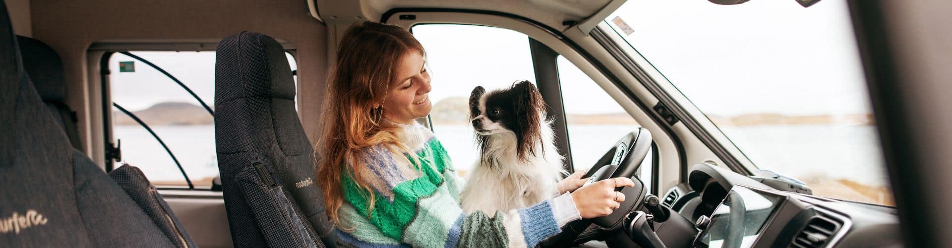 Woman with a small dog on her lap in the driver's seat of a parked camper van