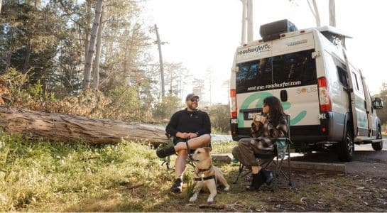 Couple relaxing in camp chairs with their dog beside a roadsurfer RV at a forest camping spot