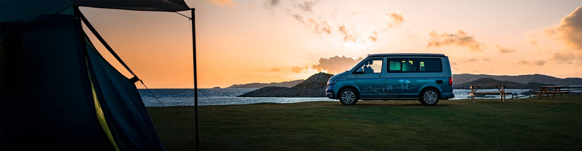 Campsite at sunset near the ocean with a tent in the foreground and a roadsurfer campervan and some picnic tables in the background