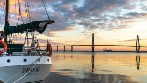 A sailboat docked at sunset with the Arthur Ravenel Jr. Bridge in the background, reflecting across Charleston Harbor.