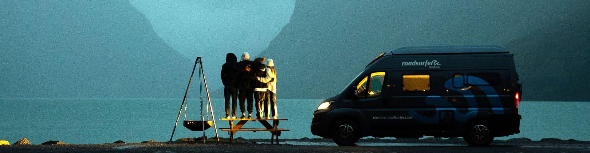 Four friends standing on a picnic table by a fjord at dusk next to a roadsurfer camper van