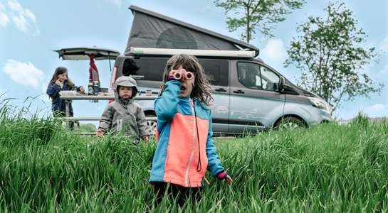 Three children playing on a green grass field with a Ford Nugget campervan standing in the background