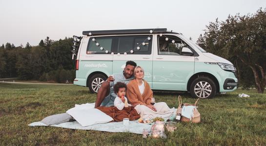 Family enjoying a picnic on the grass beside a Class B RV rental parked in a sunny field.