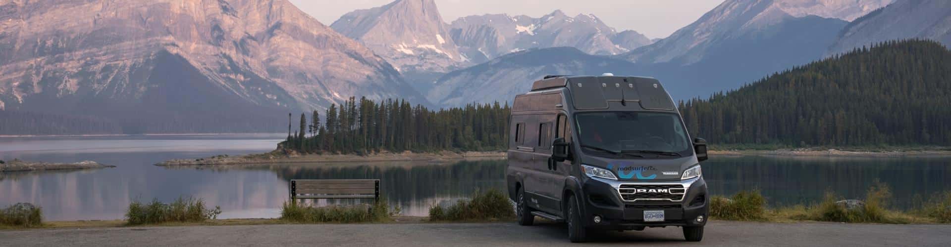 Class B RV rental parked beside a calm mountain lake with rugged peaks in the background.