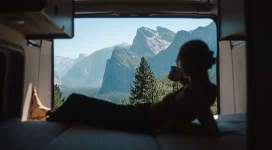 Traveler relaxing on the bed of a Class B RV rental with a panoramic mountain view through the open rear doors.