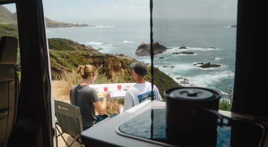 Couple eating beside their Class B RV rental overlooking dramatic ocean cliffs and rocky coastline.