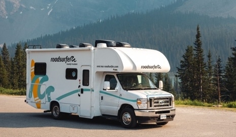A white roadsurfer Class C RV parked on a mountain road beneath towering peaks, illustrating the appeal of Class C RVs for mountain road trips.