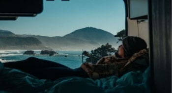 Traveler relaxing in a conversion van bed while looking out at a scenic ocean coastline during a road trip
