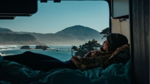 Traveler relaxing in a conversion van bed while looking out at a scenic ocean coastline during a road trip