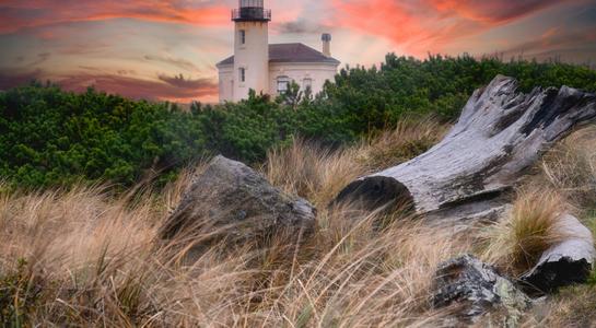 Coquille River Lighthouse in Bandon, Oregon framed by driftwood and grasses at sunset.