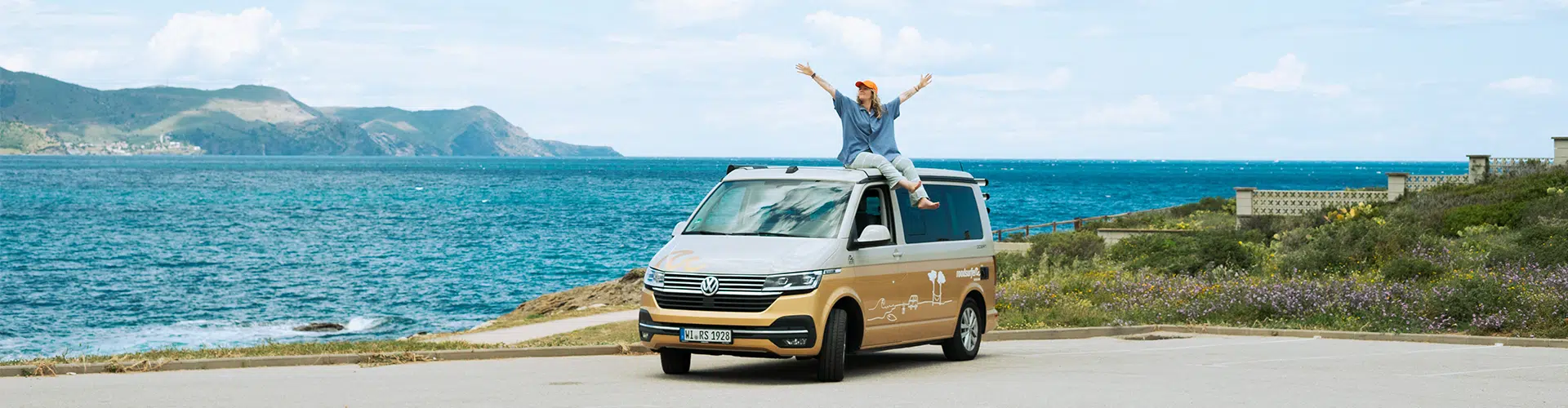 A young woman sits on her gold and white campervan parked by the Costa Brava sea, arms outstretched and cheering.