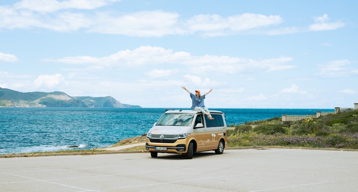 Girl sitting on the roof of a gold and white VW camper standing next to the sea at the Costa Brava in Spain