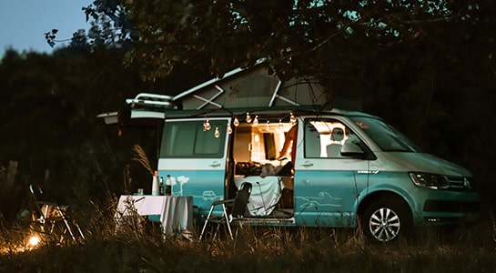 Blue campervan parked on a meadow captured during the evening with fairy lights hanging from the roof.