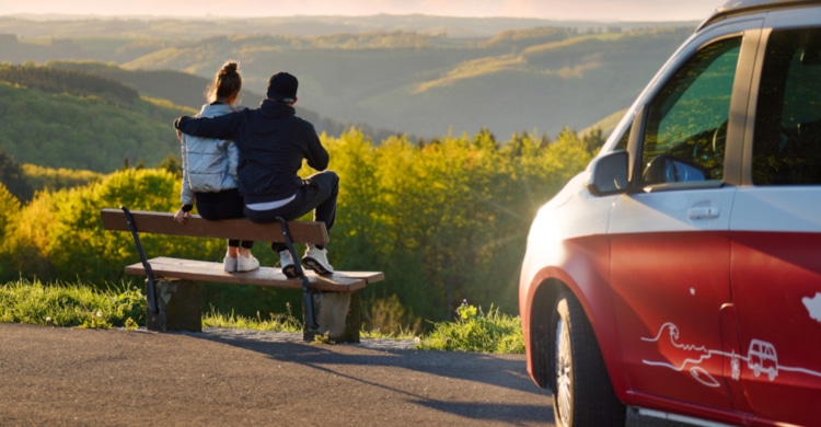 a couple sitting on a bench next to their red van, backs turned towards the camera, looking at the green hills in front of them