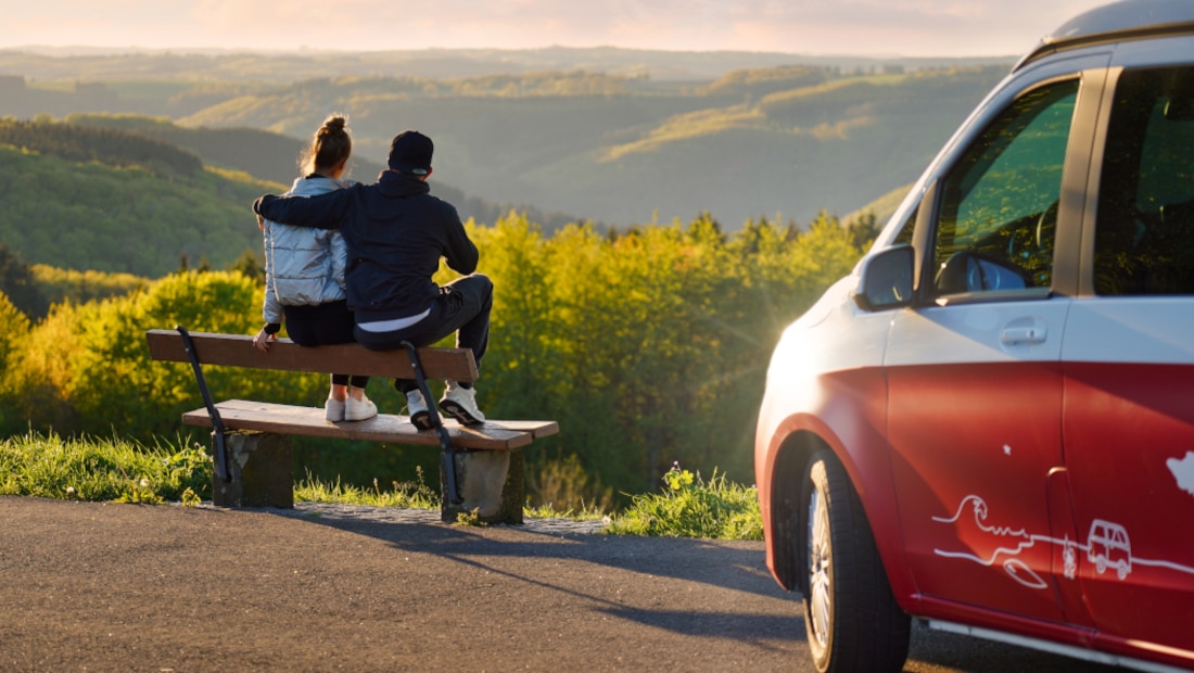 a couple sitting on a bench next to their red van, backs turned towards the camera, looking at the green hills in front of them