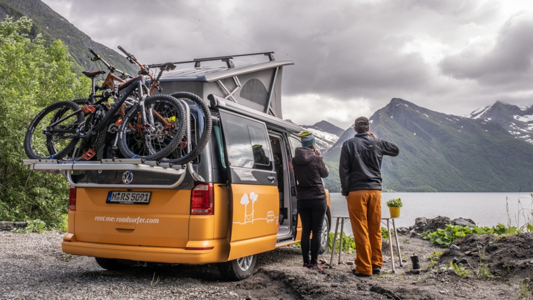 a couple standing next to a campervan with a bike rack, backs turned towards the camera, drinking coffee and looking at the mountains and the lake in front of them