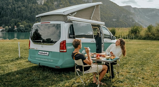 couple having breakfast next to camper van by a mountain lake