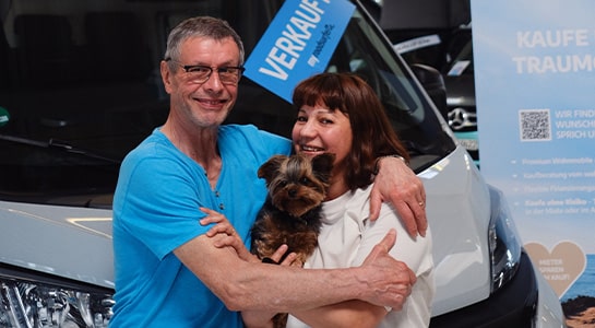 Couple hugging and holding a small dog while standing in front of a camper that shows a just-sold-sign.