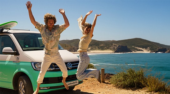 Couple jumping up next to their campervan parked at a cliff overlooking the beach.