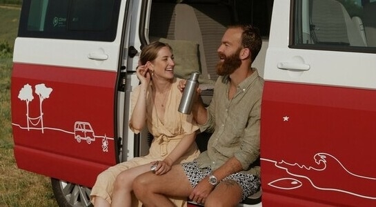 Couple Laughing While Sitting Outside Of Campervan Drinking Water