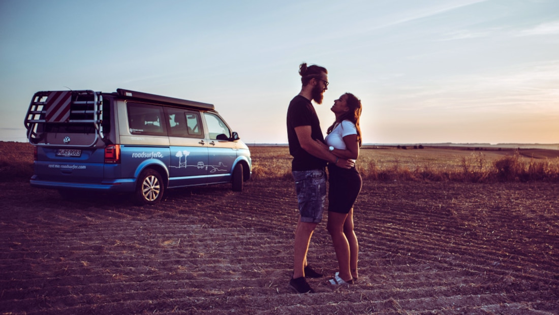 a couple standing on a field, holding and looking at each other, sunset, camper in the background