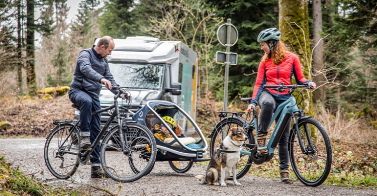 Couple sitting on bikes and with a child in a bicycle trailer. Dog is sitting next to the woman and her bike. Motorhome parked behind the couple.