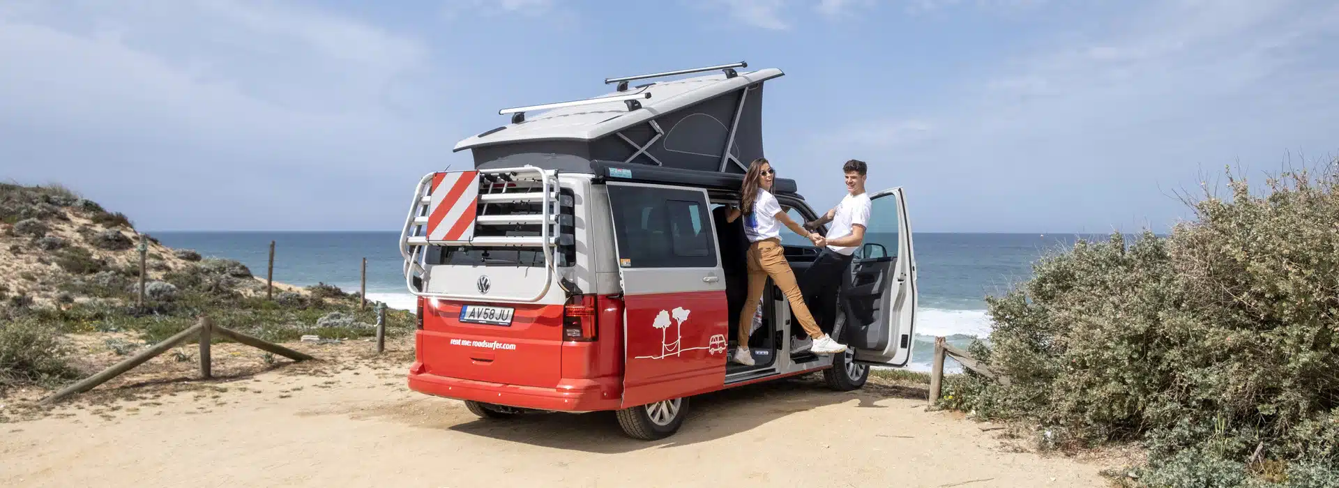 Young couple posing on a red camper van standing at the beachside with the sea in the background