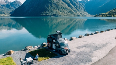 Boxvan parked at the lakeside and a couple sitting around a camping table.