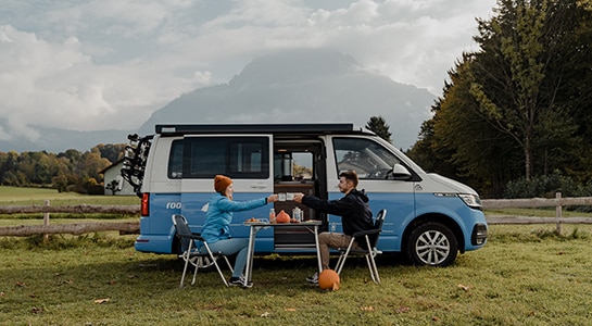 Couple sitting in front of a blue roadsurfer camper toasting with cups