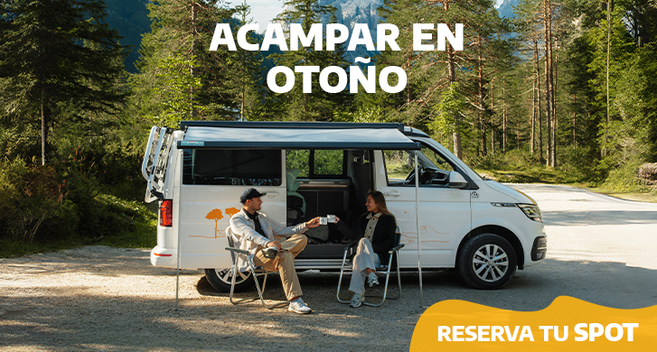 Couple sitting on camping chairs in front of their white campervan in an autumnal setting.