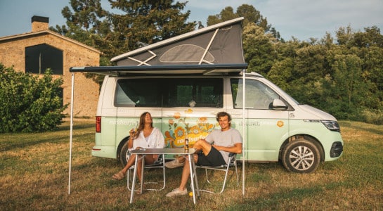 Couple sitting in the sun with drinks in front of a roadsurfer campervan on a lawn
