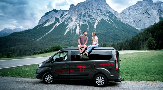 couple sitting on the roof of a camper van, mountains in the background