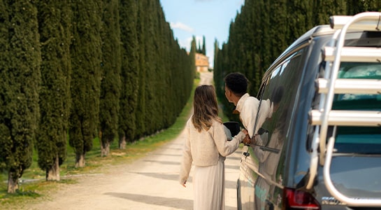 Man leaning out of window of a campervan, while woman is standing next to him. Both looking down the road.