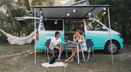 couple with dog sitting in front of a campervan in summer, hammock in the background