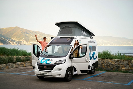 roadsurfer sprinter campervan parked in a parking lot near water and mountains with happy couple leaning out of driver and passenger doors looking at the camera