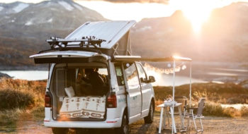 campervan facing a lake and mountains, open trunk with cozy blanket, campingtable and fairy lights on the open awning
