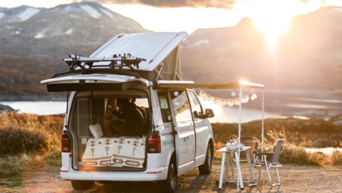campervan facing a lake and mountains, open trunk with cozy blanket, campingtable and fairy lights on the open awning