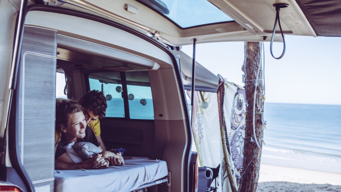 couple lying in the trunk of their van, smiling and cuddling, sea and beach in the background