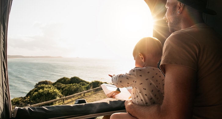 A dad holding his baby while enjoying a scenic view of the ocean and hills from the inside of a camper van during sunset.