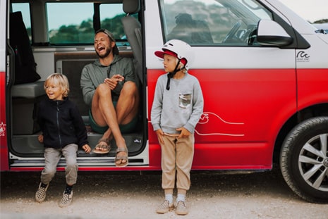 A father laughing and sitting in the doorway of a red campervan, his two sons standing next to him