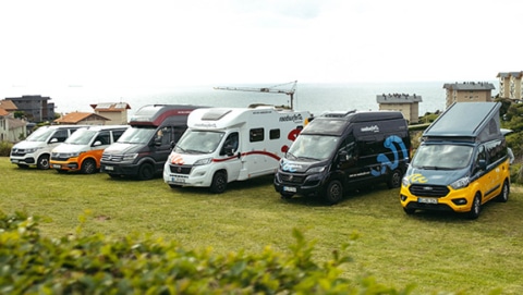 A lineup of different roadsurfer campervans and motorhomes parked on a grassy coastal field overlooking the sea, showcasing various camper models.