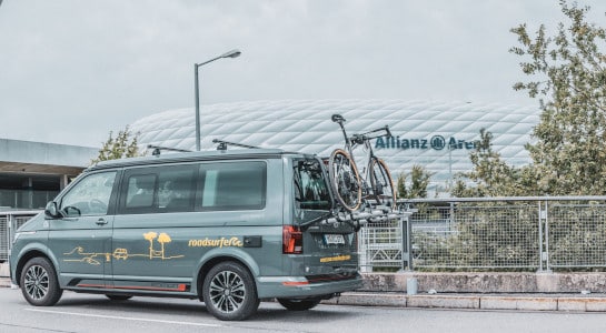 A grey campervan with a bicycle rack driving on a street, soccer stadium in the background