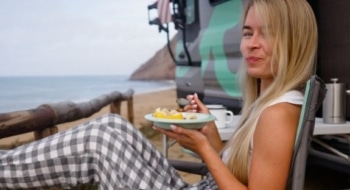 Woman eating a fresh fruit breakfast in a camp chair beside a campervan overlooking the beach, easy camping recipes lifestyle photo