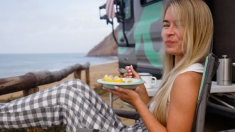 Woman eating a fresh fruit breakfast in a camp chair beside a campervan overlooking the beach, easy camping recipes lifestyle photo