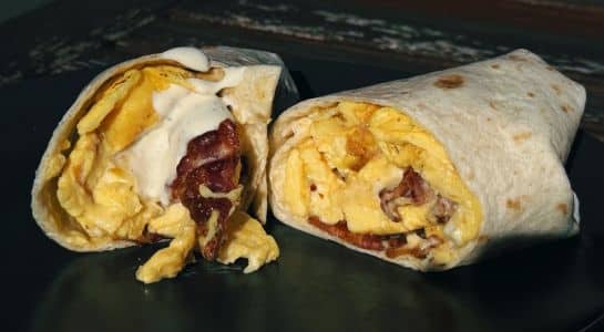 Easy camping recipes breakfast burrito sliced in half, filled with scrambled eggs, bacon, and creamy sauce, served on a dark plate