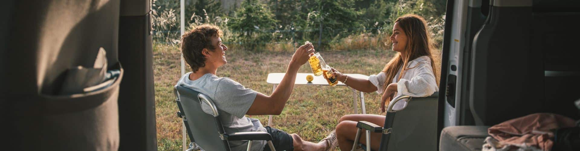 Couple sitting outside a campervan toasting with drinks at sunset, part of an easy camping recipes camping food guide