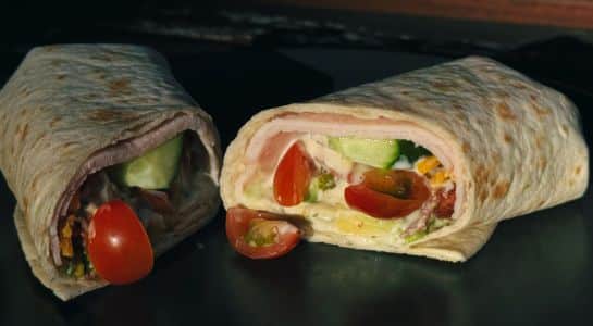 Easy camping recipes turkey wrap sliced in half, filled with turkey, cucumber, cherry tomatoes, and creamy dressing on a dark plate